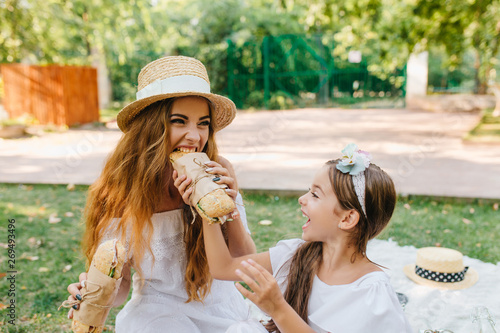 Excited girl with dark long hair looking how your mother eat bread during picnic. Hungry woman funny posing with sandwiches while her daughter laughing.