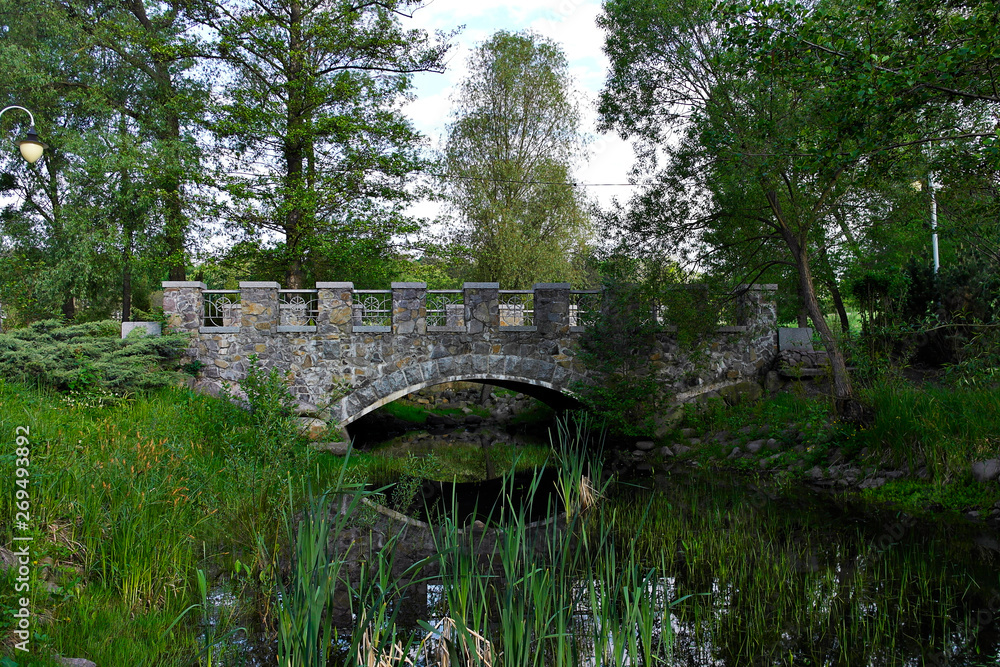 Stone bridge over the creek. flowing among large green trees and bushes. Early summer morning. Stone bridge over the creek. flowing among large green trees and bushes. Early summer morning.