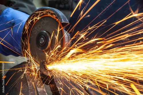 Close-up on the sides fly bright sparks from the angle grinder machine. A young male welder in a white working gloves grinds a metal product with angle grinder in the garage photo
