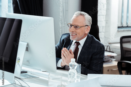 selective focus of happy businessman in glasses gesturing near computer monitor in modern office