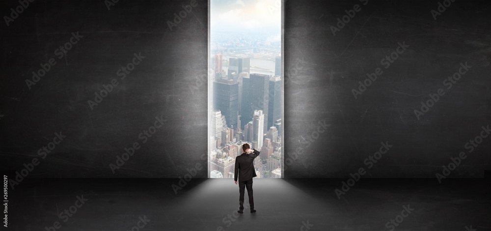 Businessman standing in a dark room and looking outside to a cityscape view
