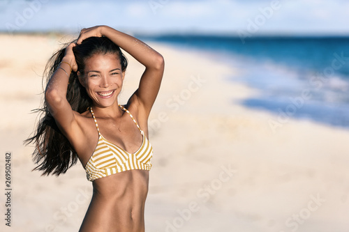 Happy beach sexy bikini body woman smiling at camera feeling confident in slim weight loss shape and toned stomach abs in swimwear and comfortable beachwear at sunset on tropical travel vacations.