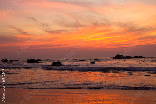 sandy beach on a background of sea waves with rocks under bright orange clouds in a purple sunset sky