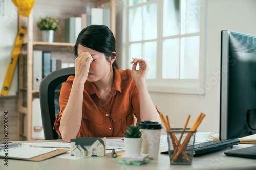 Stressed overwork and deadline. Tired young asian woman architect freelancer massaging nose bridge having headache while working in big architectural project at home office feeling exhausted sleepy photo