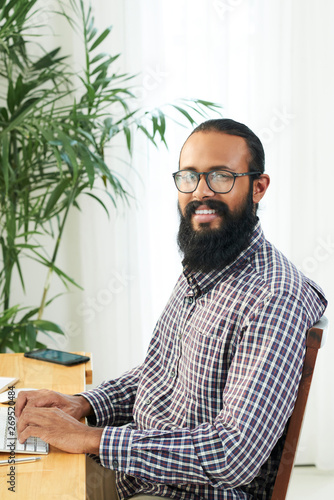 Portrait of bearded businessman in casual clothing and in eyeglasses typing on computer keyboard and smiling at camera at office