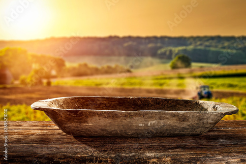 A wooden old destroyed bowl with space for your decoration. A large wooden table. Sunset in the countryside. Tractor cultivating the field. Summer time.