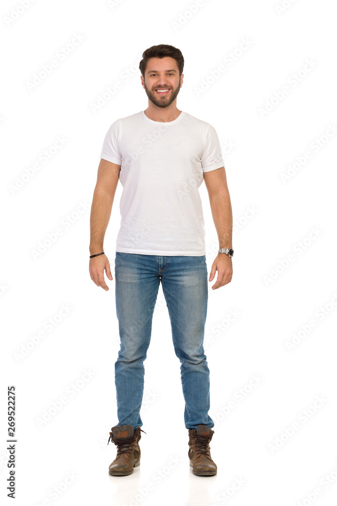 Smiling Man In Jeans And White T-shirt Is Standing And Looking At camera. Front View. Smiling Man In Jeans And White T-shirt Is Standing And Looking At camera. Front View.