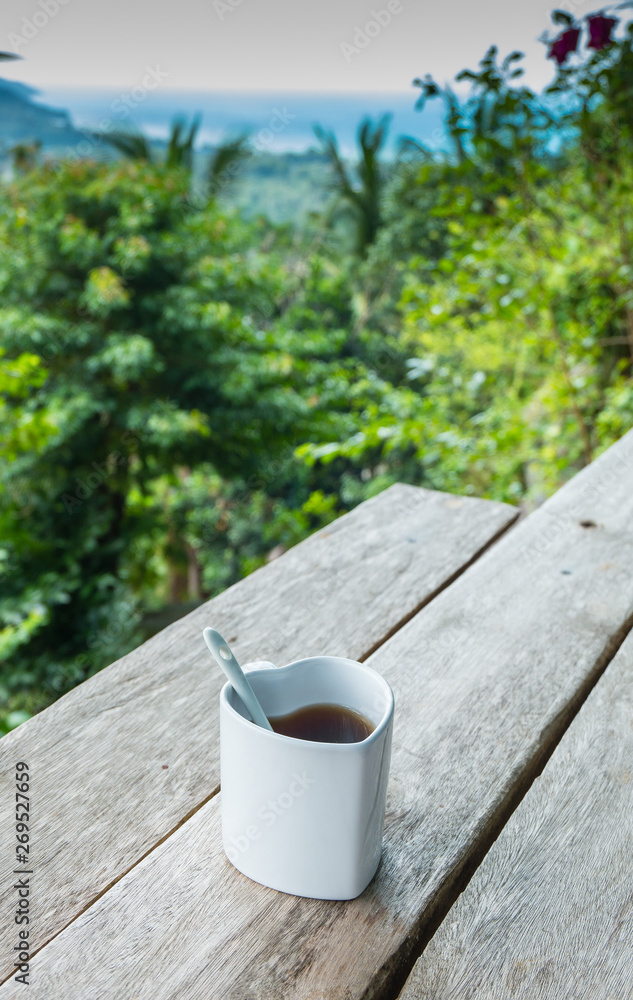 Obraz premium White coffee hot cup morning breakfast on wooden table with blurred tropical nature background. Breakfast with a Cup of coffee in the beautiful garden in nature of Phangan island, Thailand