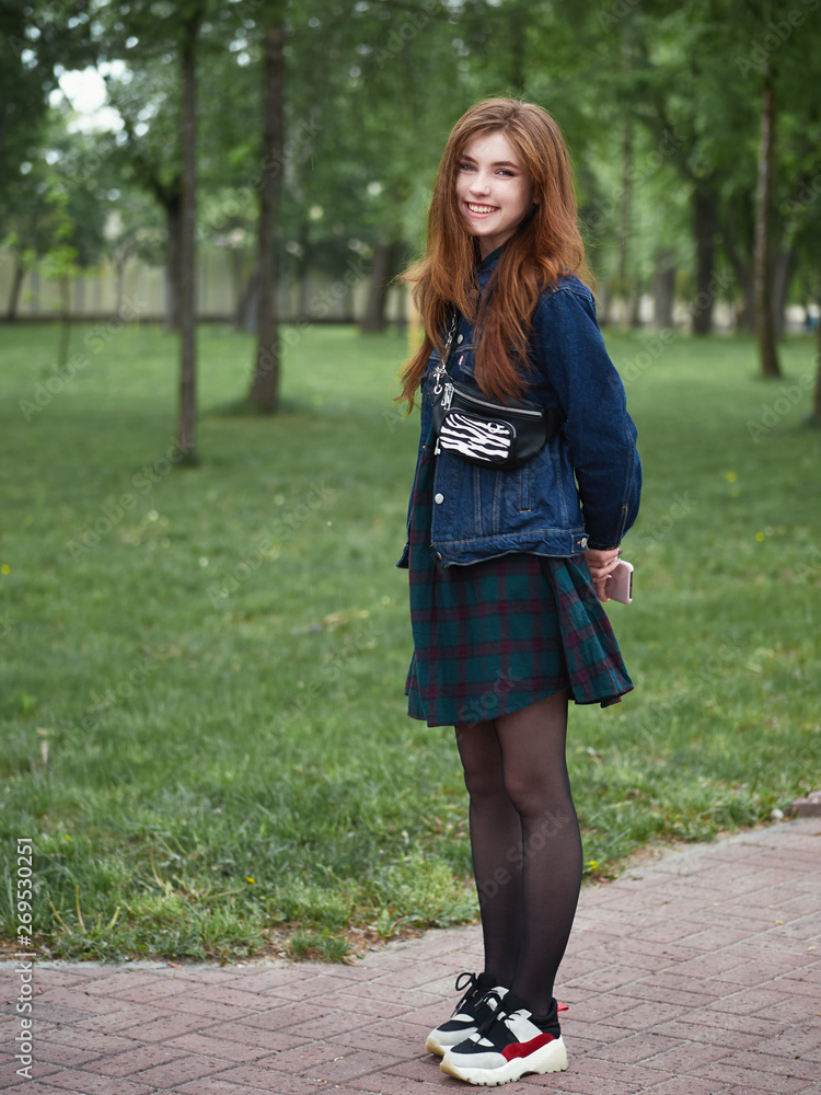 Redhead beautiful young hipster woman in stylish jeans jacket trendy plaid skirt with fashionable belt bag white sneakers walks on cloudy day with green grass city park background Redhead beautiful young hipster woman in stylish jeans jacket trendy plaid skirt with fashionable belt bag white sneakers walks on cloudy day with green grass city park background