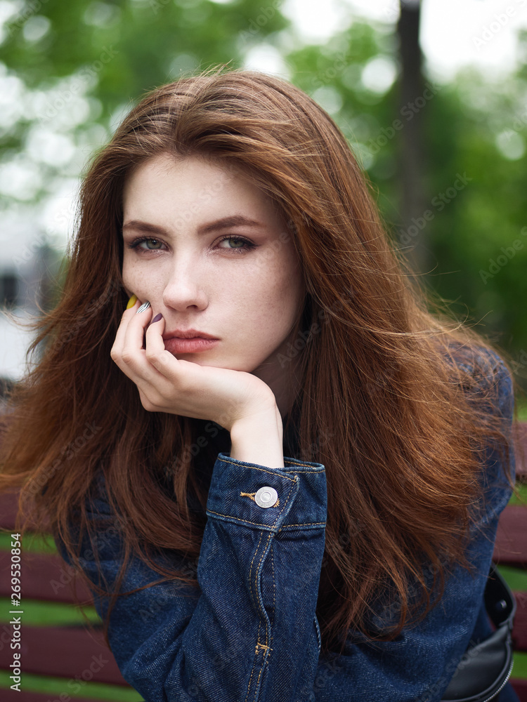 Emotional closeup portrait of naturally beautiful redhead young hipster woman in stylish jeans jacket looking into camera leaned her chin on the palm enjoying her free time on cloudy day with green Emotional closeup portrait of naturally beautiful redhead young hipster woman in stylish jeans jacket looking into camera leaned her chin on the palm enjoying her free time on cloudy day with green