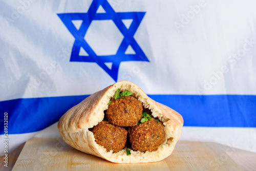 Falafel balls in a pita on wooden table over Israel flag background. Falafel plays an iconic role in Israeli cuisine and is widely considered to be the national dish of the country.