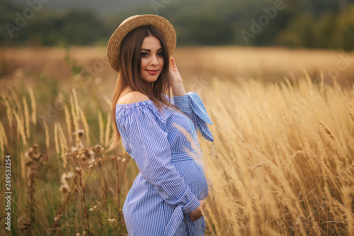 Tender pregnant woman walk in a field. Lady with hat in blue dress. Relax time. Woman is waiting for a baby