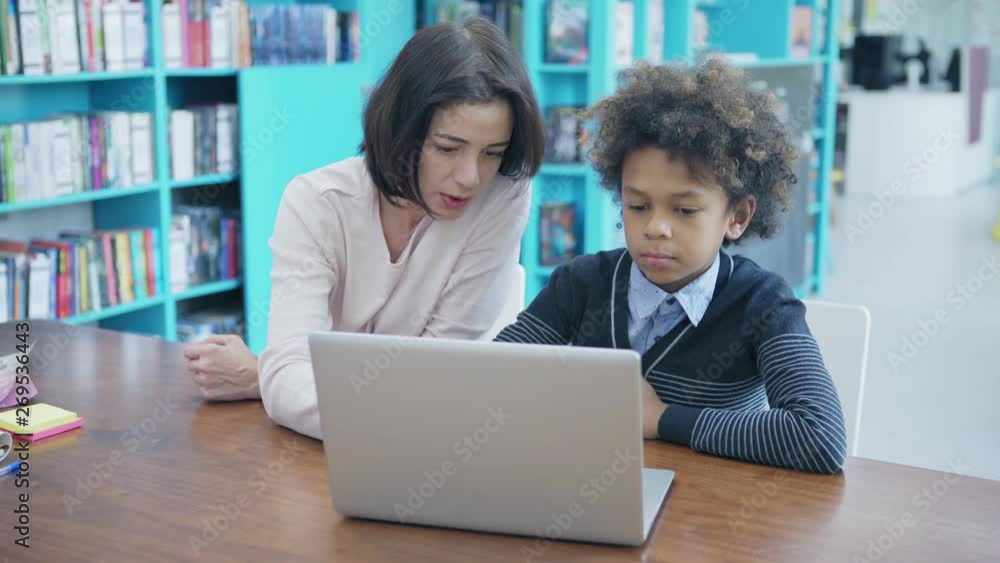 Female teacher and elementary mixed race student with curly hair sitting at desk in classroom and using laptop together, tracking medium shot Female teacher and elementary mixed race student with curly hair sitting at desk in classroom and using laptop together, tracking medium shot