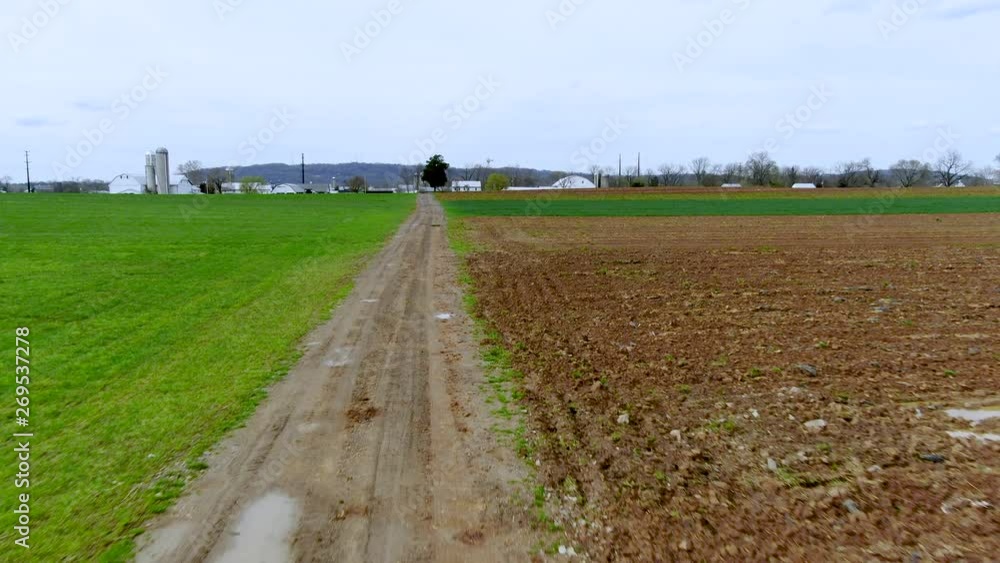 Flying above muddy dirt road in rural countryside, small village with farm, silos and barns in background, fields and meadows at both sides of the road, agriculture and farming concept Flying above muddy dirt road in rural countryside, small village with farm, silos and barns in background, fields and meadows at both sides of the road, agriculture and farming concept
