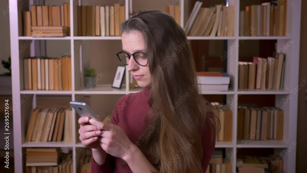 Closeup portrait of young caucasian female student in glasses using the phone smiling happily looking at camera in the college library indoors Closeup portrait of young caucasian female student in glasses using the phone smiling happily looking at camera in the college library indoors
