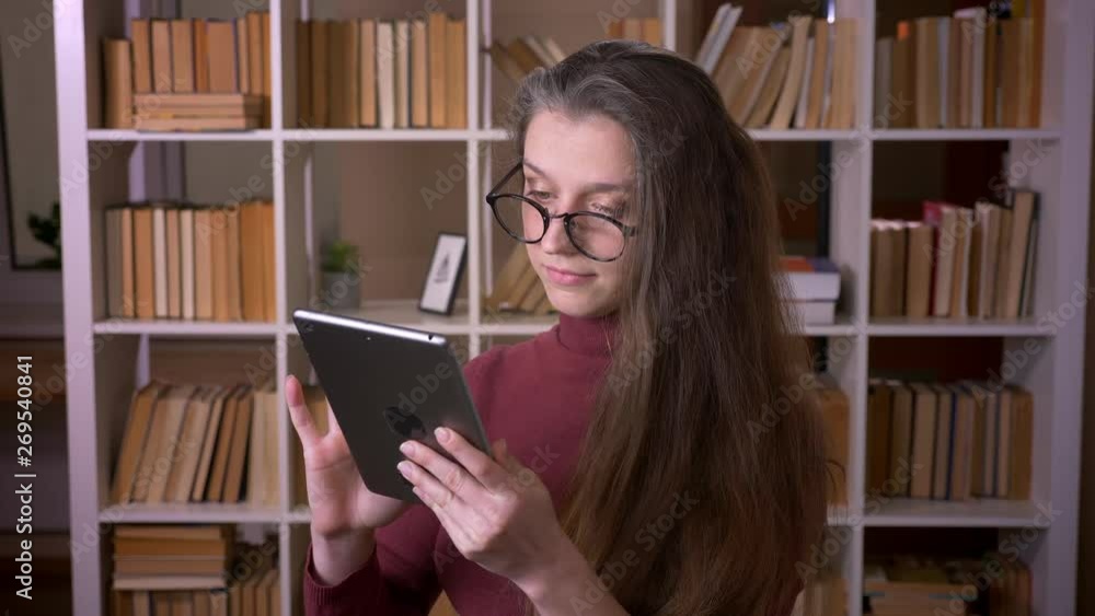 Closeup portrait of young caucasian female student in glasses typing on the tablet indoors in the college library indoors Closeup portrait of young caucasian female student in glasses typing on the tablet indoors in the college library indoors