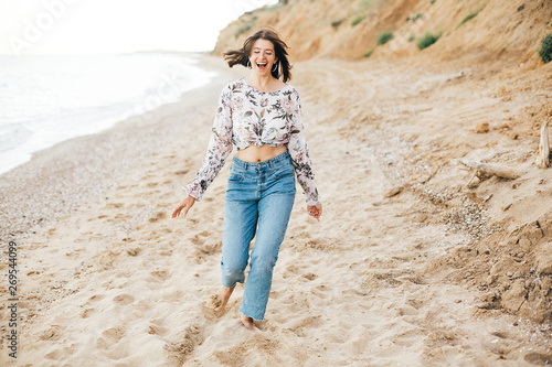 Stylish hipster girl running on beach at sea and smiling. Happy boho woman in denim jeans and floral blouse relaxing at sandy cliff on tropical island. Travel and summer vacation concept © sonyachny