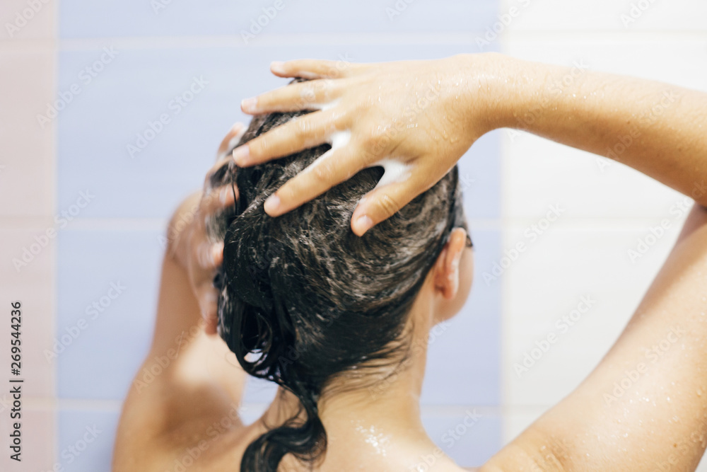 Naklejka premium Young happy woman washing her hair with shampoo, hands with foam closeup. Back of beautiful brunette girl taking shower and enjoying relax time. Body and hair hygiene, lifestyle concept