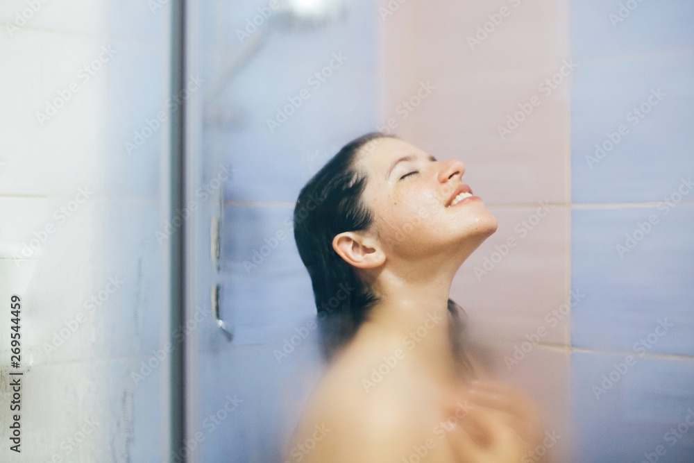 Young happy woman taking hot shower at home or hotel bathroom. Sensual portrait of beautiful brunette girl enjoying time in shower. Body and skin hygiene, lifestyle concept. Space text