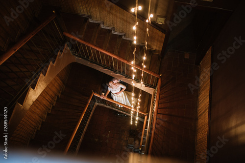 The groom in a suit and the bride in a wedding dress are standing on a wooden staircase