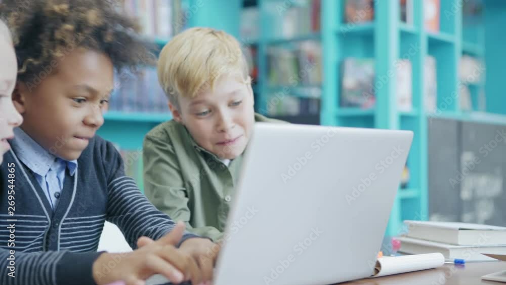 Tracking right medium shot of three curious diverse students with laptop sitting at desk in elementary school classroom. Mixed  race schoolboy playing computer game, his friends supporting him