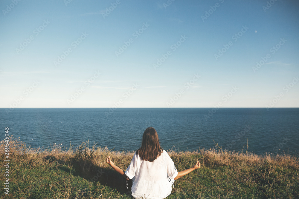 Yoga day. Stylish hipster girl sitting in yoga pose and relaxing on mountain with sea view. Happy boho woman practicing yoga and meditation on tropical island. Mindful concept. Space for text Yoga day. Stylish hipster girl sitting in yoga pose and relaxing on mountain with sea view. Happy boho woman practicing yoga and meditation on tropical island. Mindful concept. Space for text