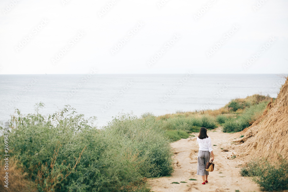 Stylish hipster girl relaxing on beach.  Happy young boho woman in white shirt walking on tropical island at sandy cliff and grass. Summer vacation. Space for text