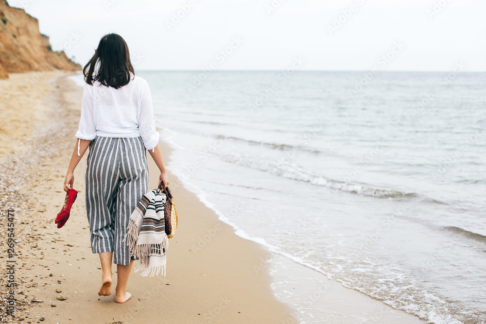 Φωτογραφία Stylish hipster girl walking barefoot on beach, holding bag and shoes in hand