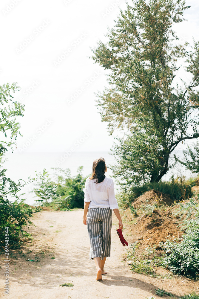 Stylish hipster girl relaxing on beach. Happy young boho woman in white shirt walking barefoot on tropical island at sandy cliff and trees. Summer vacation. Space for text Stylish hipster girl relaxing on beach. Happy young boho woman in white shirt walking barefoot on tropical island at sandy cliff and trees. Summer vacation. Space for text