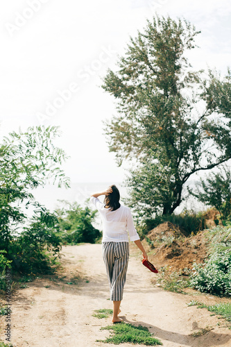 Happy young boho woman in white shirt walking barefoot on tropical island at sandy cliff and trees. Summer vacation. Space for text. Stylish hipster girl relaxing in park at beach.