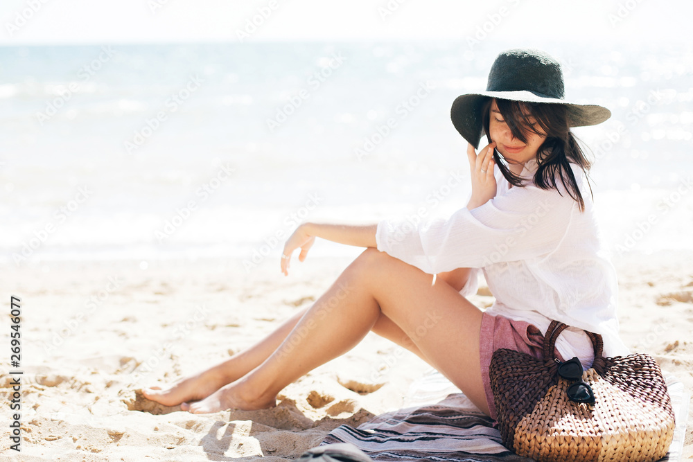 Stylish hipster girl in hat sitting on beach and tanning near sea waves. Summer vacation. Happy boho woman relaxing and enjoying sunny warm day at ocean. Space for text - obrazy, fototapety, plakaty 