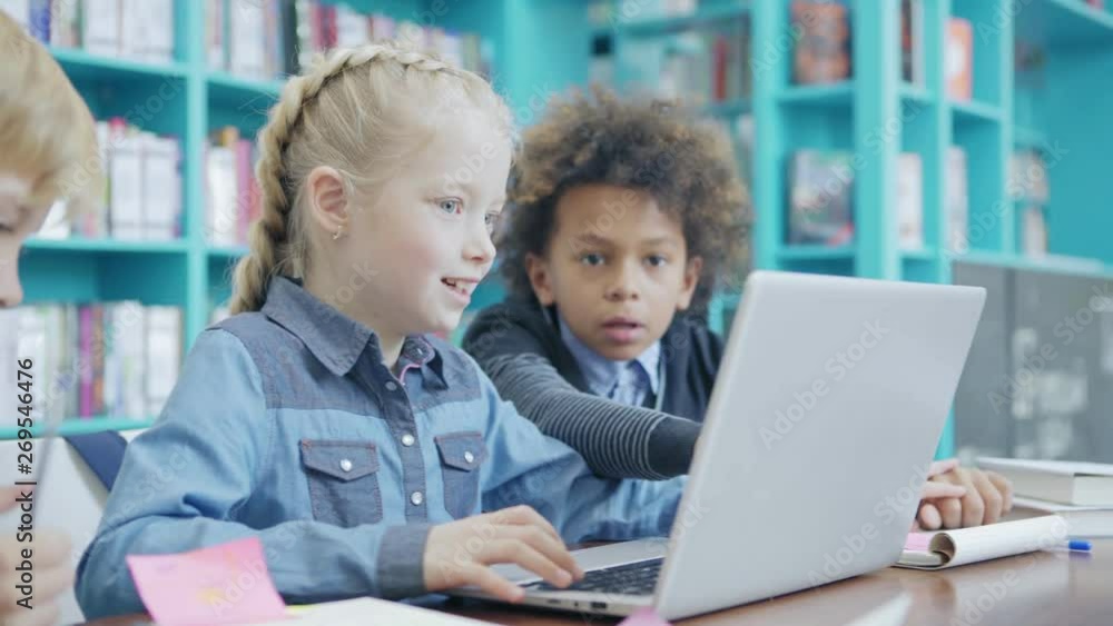 Group of three elementary students sitting at desk in classroom with laptop in front of them. Mixed race boy helping Caucasian girl to do task on computer, tracking right shot Group of three elementary students sitting at desk in classroom with laptop in front of them. Mixed race boy helping Caucasian girl to do task on computer, tracking right shot