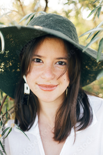 Portrait of young fashionable woman with modern earrings posing in green olive branches in soft evening light, stylish boho girl in hat relaxing on tropical island. Summer vacation