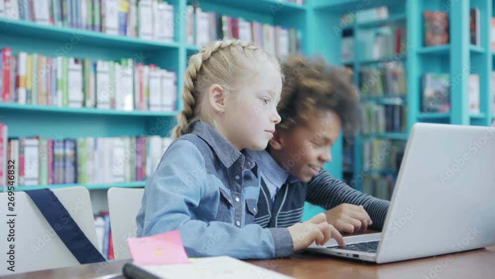 Two diverse elementary school children, boy and girl, sitting at desk in classroom and doing task or playing educational game on laptop together Two diverse elementary school children, boy and girl, sitting at desk in classroom and doing task or playing educational game on laptop together