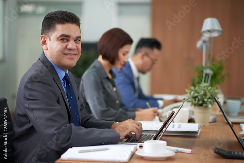 Portrait of mature businessman typing on laptop computer and looking at camera while sitting at business meeting together with his colleagues at office