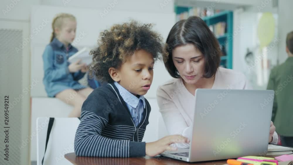 Rack focus of elementary school girl reading textbook while teacher and mixed race boy discussing task on laptop computer sitting at desk in elementary school classroom Rack focus of elementary school girl reading textbook while teacher and mixed race boy discussing task on laptop computer sitting at desk in elementary school classroom