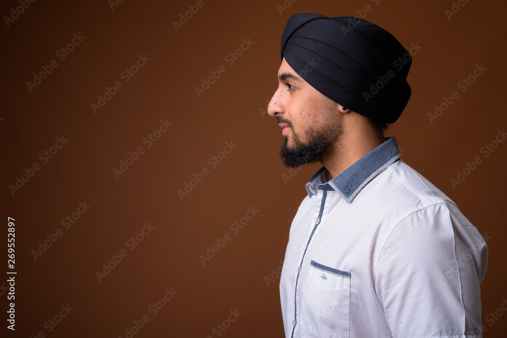 Young bearded Indian Sikh man wearing turban against brown backg Young bearded Indian Sikh man wearing turban against brown backg