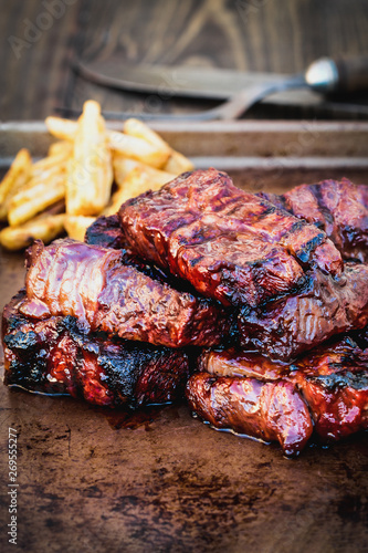 Bbq boneless beef ribs with barbecue sauce and potato wedges over a rustic background. Extreme shallow depth of field with blurred background and selective focus on front of meat