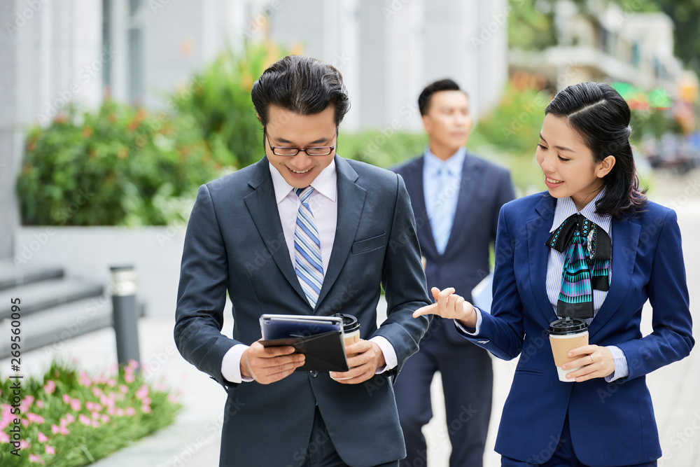 Smiling young businesswoman drinking coffee and discussing online work with her colleague while he using digital tablet outdoors Smiling young businesswoman drinking coffee and discussing online work with her colleague while he using digital tablet outdoors