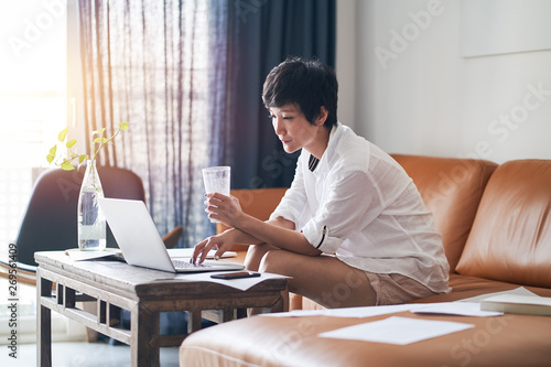 Photography Asian self employed woman sitting on couch  drinking water & working on laptop a