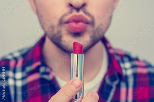 Man bearded, metrosexual holding red lipstick in his hand close to face, on camera, front view, close up, cropped image, toned