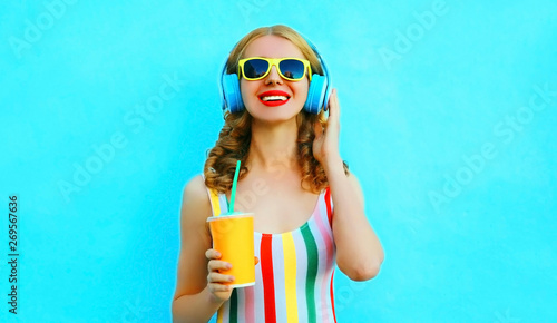 Happy smiling woman holding cup of juice listening to music in wireless headphones on colorful blue background
