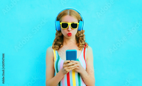 cool girl blowing red lips holding phone listening to music in wireless headphones on colorful blue background © rohappy