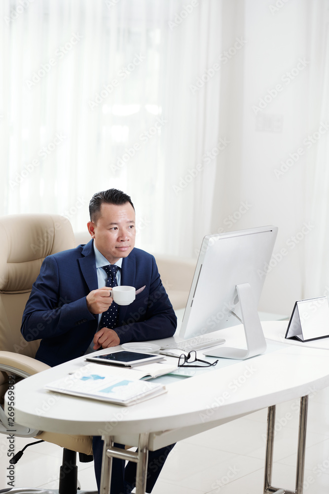 Asian mature businessman sitting at the table looking at computer monitor holding cup and drinking coffee at office