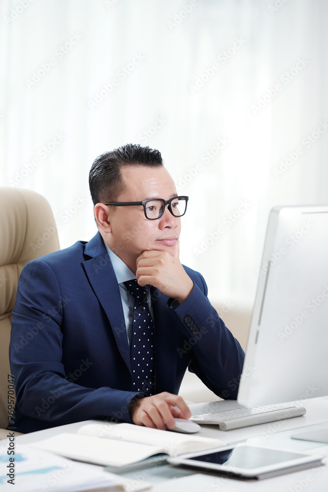 Serious confident businessman wearing eyeglasses working on computer while sitting at desk at office Serious confident businessman wearing eyeglasses working on computer while sitting at desk at office