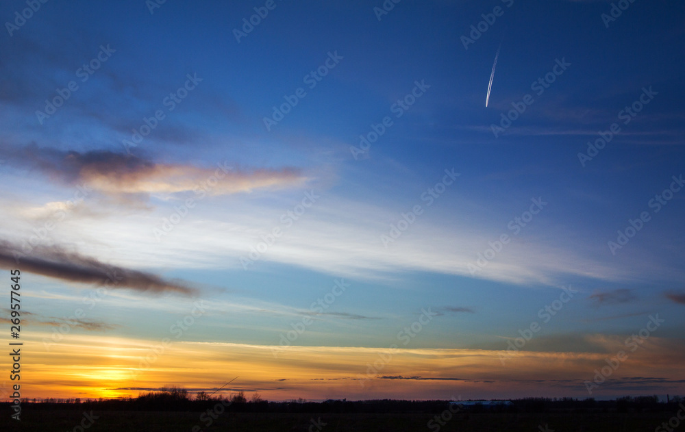 Sunset, sunlight from behind the horizon. High in the evening sky the plane flies, leaving a white trail on the blue sky.