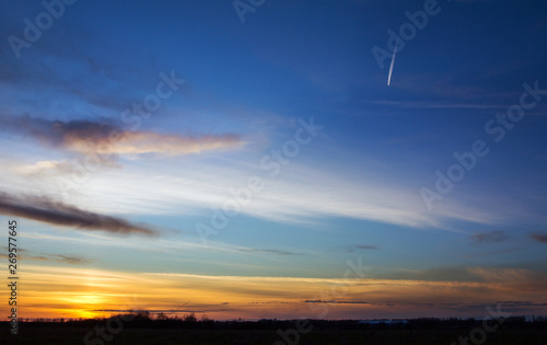 Sunset, sunlight from behind the horizon. High in the evening sky the plane flies, leaving a white trail on the blue sky.
