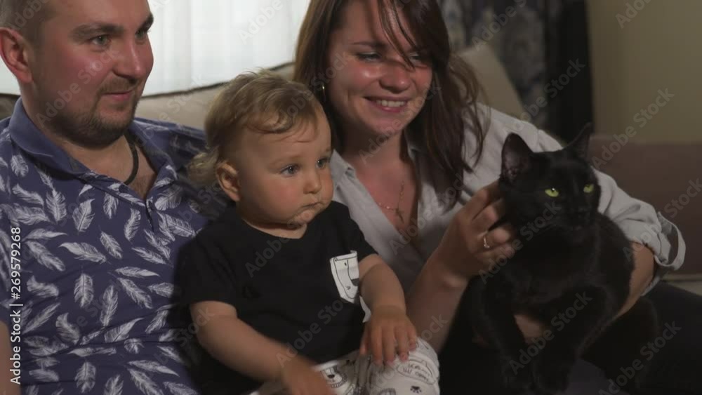 Family values: Mother and father playing with baby child boy indoor - Huge black panther like cat kitty - Parents wearing blue and gray home wear and their son black and white - Happy smiling people Family values: Mother and father playing with baby child boy indoor - Huge black panther like cat kitty - Parents wearing blue and gray home wear and their son black and white - Happy smiling people