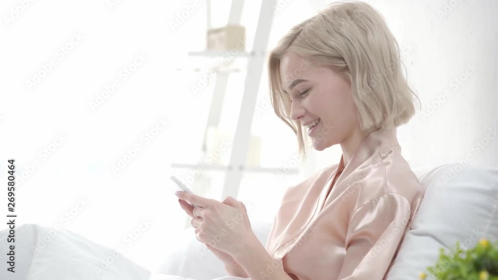 beautiful blonde girl using smartphone, smiling, covering mouth and laughing in bedroom beautiful blonde girl using smartphone, smiling, covering mouth and laughing in bedroom