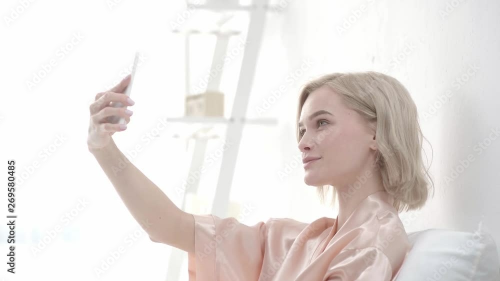 cheerful blonde girl taking selfie on smartphone, smiling, sending kisses and touching face at home cheerful blonde girl taking selfie on smartphone, smiling, sending kisses and touching face at home